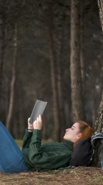 Kızıl saçlı genç kadın ormanda ağacın altında kitap okuyor. Doğada dinlenmek..
