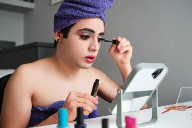 Young drag queen applying eye mascara after shower with a towel on his head.