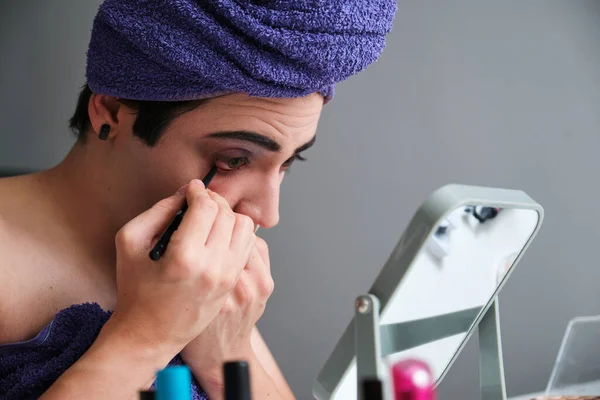 Young transgender man applying eyeliner after shower with a towel on his head.