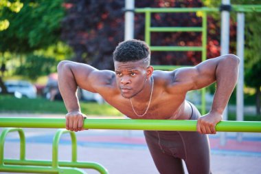 Young fit shirtless black man doing push-ups on a bar in a calisthenics park outdoors on sunny day. Fitness and sport lifestyle.