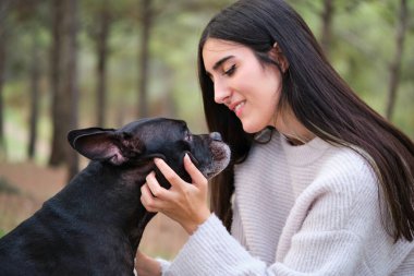 Beyaz bir kadın, Amerikan personeli ile Fransız bulldog köpeğini çam ormanında okşuyor..