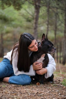 Kafkasyalı genç bir kadın çam ormanında Amerikan personeliyle Fransız bulldog köpeğine sarılıyor..