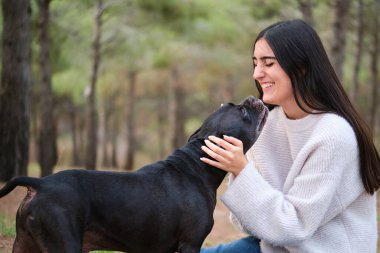 Amerikan personeli ile Fransız buldoğunun karışımı melez köpeğinin ardından gülen beyaz bir kadın çam ormanında onu öpüyor..