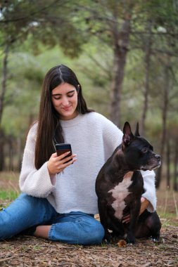Kafkasyalı genç bir kadın Amerikan Stadyumu ve Fransız buldogu ile akıllı telefon kullanıyor. Çam ormanında oturan melez bir köpek..