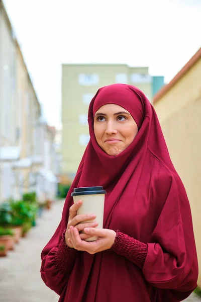 Portrait of muslim young woman in hijab shrugging holding a coffee cup in the street.