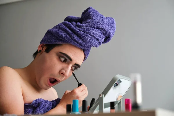 Young gender fluid person applying eye mascara after shower with a towel on his head.