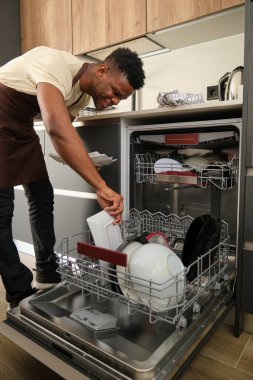 Young african man putting dishes in the dishwasher in a kitchen.