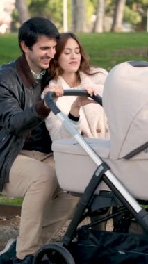 Couple looking at their baby while rocking the stroller playing with their dog sitting on a bench in a park.