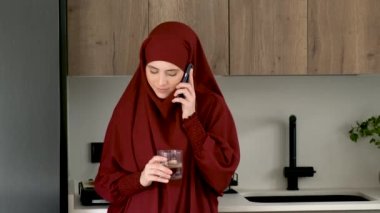 Başörtüsü takmış, su içen ve evde, mutfakta akıllı telefondan konuşan Müslüman genç bir kadın..