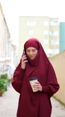 Muslim young woman in hijab talking on smartphone and holding a coffee cup in the street.