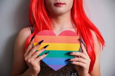 Unrecognizable transgender person holding a rainbow flag heart on grey background. LGBTQ queer.