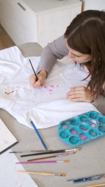 Top view of a smiling caucasian woman painting a design in a white shirt. Womans creative expression through painted clothing designs.