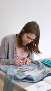 Caucasian woman hand painting a design using acrylic paints of a deer and flowers in a denim jacket. Unique clothing designs.