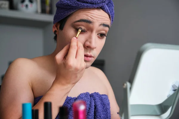 Young transgender man applying eyeshadow after shower with a towel on his head.