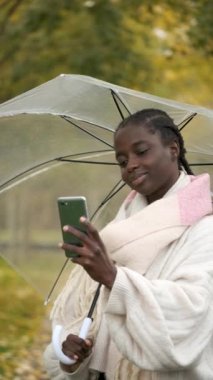 Sonbaharda gülümseyen akıllı telefon ve şeffaf şemsiyeyle selfie çeken Afrikalı bir kadın..