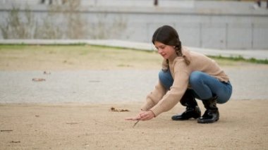 Girl with two braids drawing hopscotch in the sand with a stick outdoors.