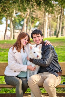 Genç beyaz bir çift parktaki bir bankta köpeğini okşuyor. Aile.