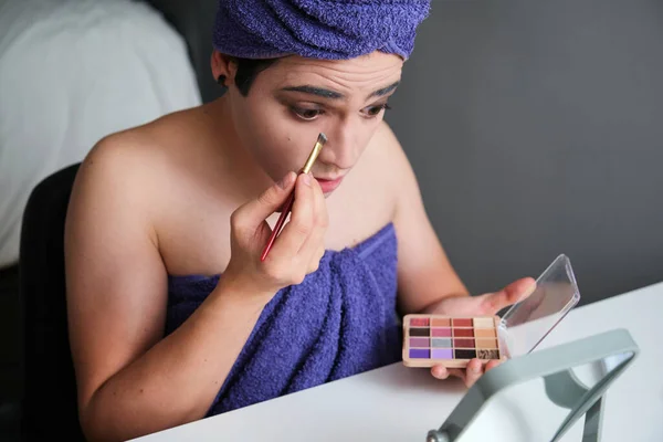 Young gender fluid person man applying eyeshadow after shower with a towel on his head.
