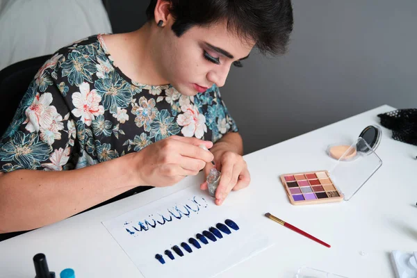 Young transgender man in a dress painting false nails with glitter nail polish.