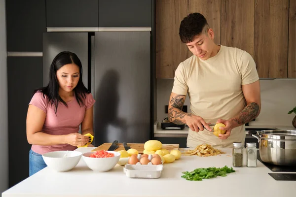 Two young latin people peeling potatoes and cleaning kitchen counter to prepare a recipe at kitchen.