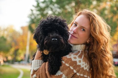 Young woman hugging her black puppy poodle dog in a park in autumn.