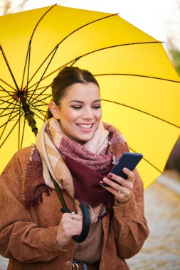 Sonbaharda telefonu kullanan ve elinde sarı bir şemsiye tutan beyaz bir kadın gülümsüyor..