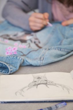 Tanımlanamayan bir kadın, kot ceketli bir geyik ve çiçeklerin akrilik boyalarıyla bir tasarım yapıyor. Benzersiz kıyafet tasarımları.