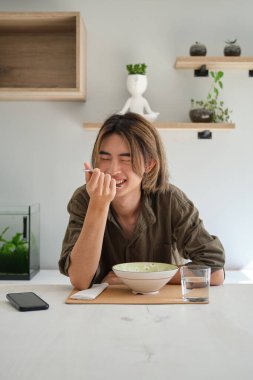 Smiling asian young man eating soup in the living room. Having lunch.