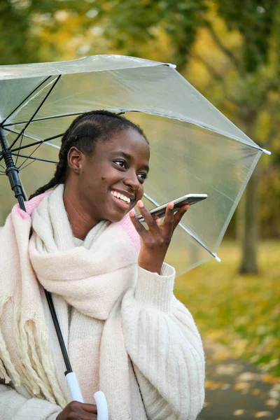 stock image African woman with a transparent umbrella smilling and recording audio message in autumn.