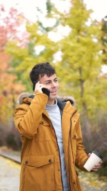 Gülümseyen genç adam akıllı telefondan konuşuyor ve bir sonbahar günü elinde kahve tutuyor..