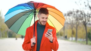 Sonbaharda sokakta telefon kullanan kırmızı paltolu ve gökkuşağı şemsiyeli Latin bir genç..