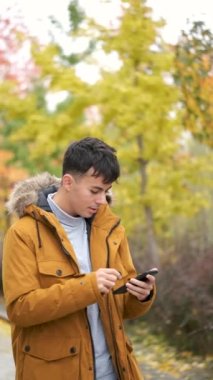 Rüzgarlı bir sonbahar gününde akıllı telefon kullanan gülümseyen genç adam..
