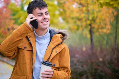 Gülümseyen genç adam akıllı telefondan konuşuyor ve bir sonbahar günü elinde kahve tutuyor..