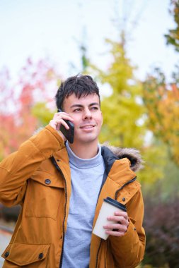 Gülümseyen genç adam akıllı telefondan konuşuyor ve bir sonbahar günü elinde kahve tutuyor..