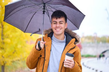 Yağmurlu bir sonbahar gününde sokakta elinde kahve ve şemsiye tutan gülümseyen genç bir adamın portresi..