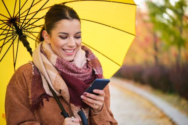 Sonbaharda telefonu kullanan ve elinde sarı bir şemsiye tutan beyaz bir kadın gülümsüyor..