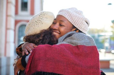 İki çok kültürlü kadın mutlu arkadaş birbirine sarılıyor ve caddede gülümsüyor..