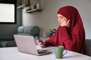 Evde çevrimiçi bir dersteyken tesettürlü genç Müslüman kız üniversite öğrencisi telefon kullanıyor.