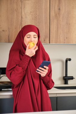 Muslim young woman in hijab drinking coffee and using smartphone in the kitchen at home.