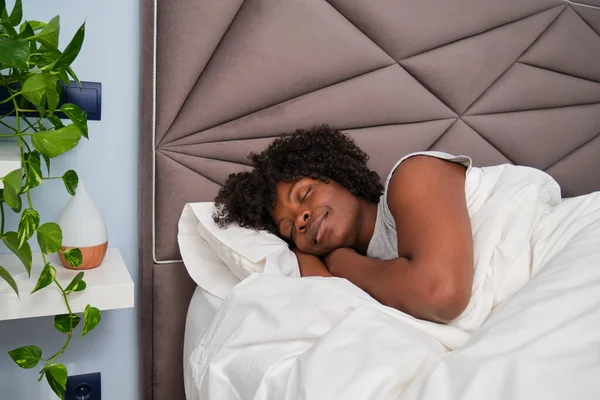 stock image Happy african young woman sleeping on white sheets in bed in a room with a plant. Resting asleep.