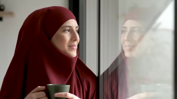 穆斯林女青年头戴头巾 拿着咖啡杯微笑着从家里的窗户往外看 — 图库视频影像