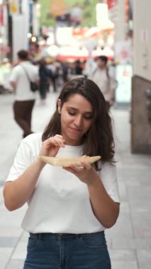 Latin kökenli genç bir kadın takoyaki yiyor ve Japonya 'nın Osaka şehrindeki Dotonbori caddesindeki kameraya bakıyor..