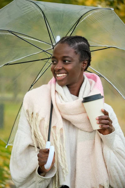 stock image African woman with a coffee and transparent umbrella smiling in autumn.