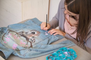 Caucasian woman hand painting a design using acrylic paints of a deer and flowers in a denim jacket. Unique clothing designs. clipart