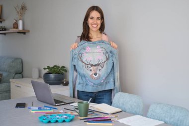 Portrait of an artist and entrepreneur woman smiling and showing her art design in a denim jacket. Unique clothing designs. clipart