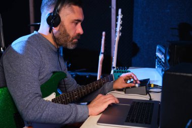Müzisyen adam ev kayıt stüdyosunda elektro gitarının sesini ekolayzır, kulaklık ve dizüstü bilgisayarla ayarlıyor. Müzik üretimi.