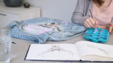Unrecognizable woman painting a design of a deer and flowers in a denim jacket. Womans creative expression through painted clothing designs.