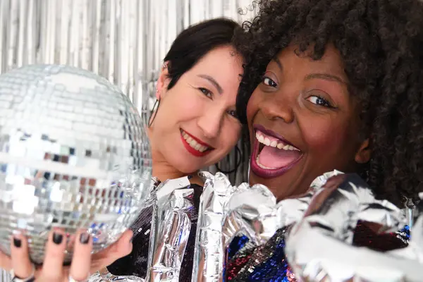 Two multiracial women in a party with a party silver balloon and disco ball taking a selfie. Two friends laughing at a party.