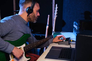 Müzisyen adam ev kayıt stüdyosunda elektro gitarının sesini ekolayzır, kulaklık ve dizüstü bilgisayarla ayarlıyor. Müzik üretimi.