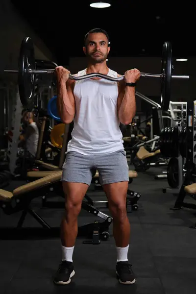 Strong Hispanic man doing barbell curl for biceps strength at a gym.
